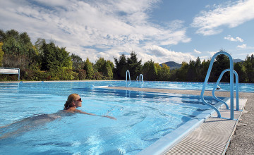Blick auf ein Schwimmbecken mit zwei Einstiegsstiegen und einer Frau, die darin schwimmt. Im Hintergrund sind Bäume.