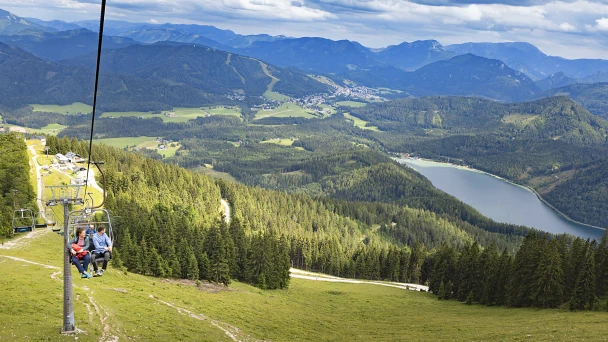 Blick auf den Zweiersessellift, in dem zwei Personen fahren. Rechts davon sieht man den Ausblick ins Tal mit einer hügeligen Wiesen- und Waldlandschaft, sowie einen See.