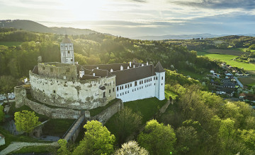 Blick auf die Schallaburg von schräg oben. Es sind alte Tormauern mit einem neuern Teil des Schlosses zu sehen. Darum herum ist eine Hügellandschaft mit Bäumen.
