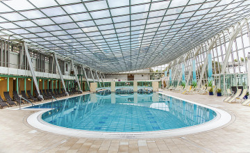 Blick auf ein ovales Schwimmbecken, dav von einem Fliesenboden und Liegestühlen umgeben ist. Die Decke und rechte Wand sind verglast.