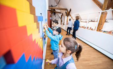 Kinder stehen vor einer bunten Wand, die interaktive Elementen und spielen. Im Hintergrund sieht man eine weiße Vitrine mit Ausstellungsobjekten.