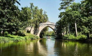 Blick auf einen Fluss, der von viel Grün umgeben ist und ein Boot, das unter einer imposanten Brücke durchfährt.