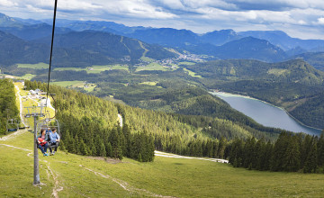Blick auf den Zweiersessellift, in dem zwei Personen fahren. Rechts davon sieht man den Ausblick ins Tal mit einer hügeligen Wiesen- und Waldlandschaft, sowie einen See.