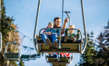Blick auf einen Zweier-Sessellift auf dem ein Vater neben einem Kind sitzt und auf etwas zeigt. Auf seinem Schoß sitzt ein Kleinkind. Im Hintergrund sieht man weitere Sessellifte und Bäume.