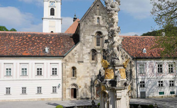 Blick auf das Stift Heiligenkreuz hinter dem sich ein Kirchenturm erhebt. Davor steht eine Gedenksäule aus Stein.