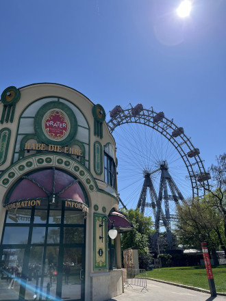 Gebäude im Vordergrund, Riesenrad im Hintergrund, blauer Himmel und Sonnenschein